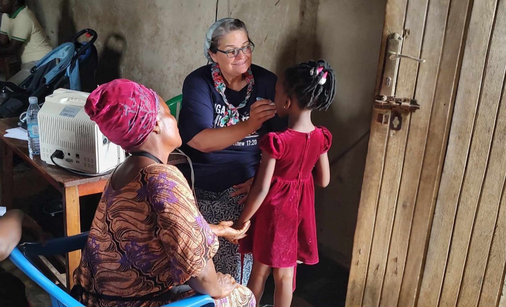 Checking a child's temperature at their home, Bonnie smiles. Presumably the child's mother is sat to the side, offering her kid support with a gentle hand on their arm. Bonnie is wearing a bible verse t-shirt while the mother and child both wear red.
